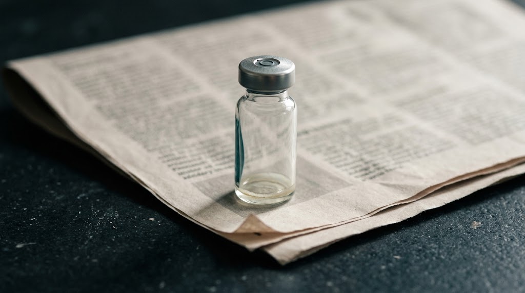 A clear glass vial standing on an open newspaper on dark stone
