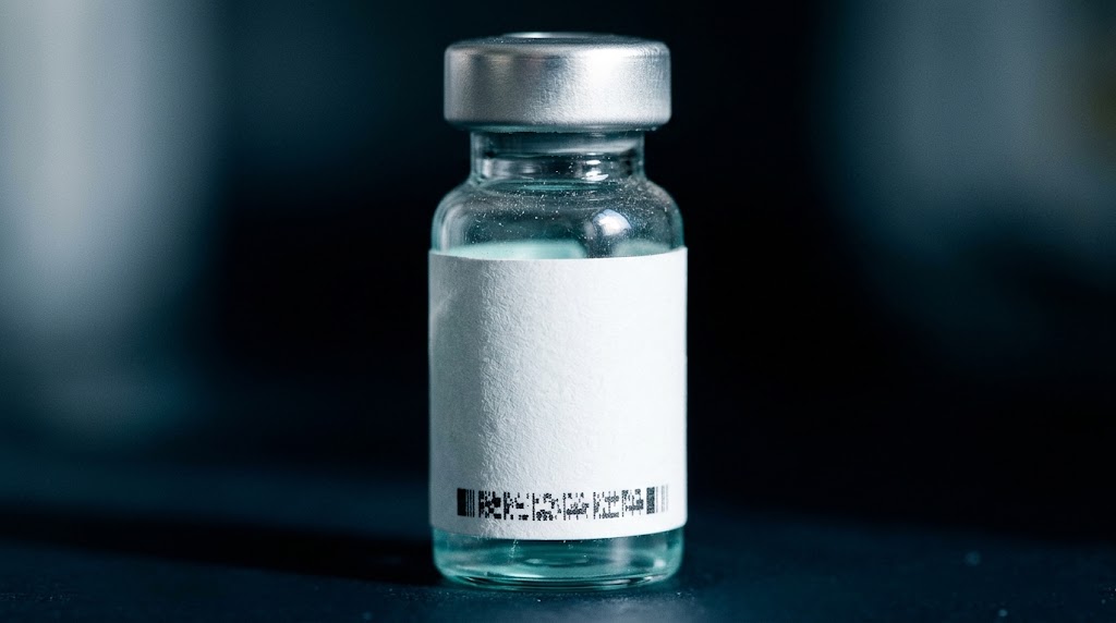 A glass peptide vial standing on a printed page of regulation text, the blank area of its label catching the light