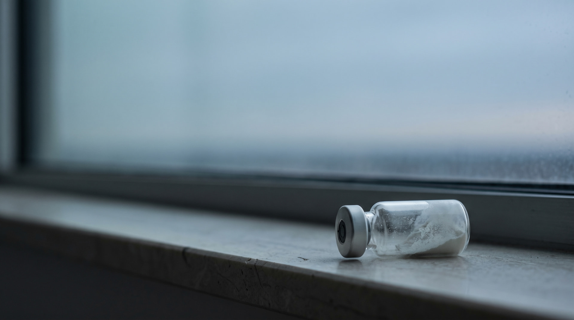 A clear research vial containing white powder on a cool, dim windowsill