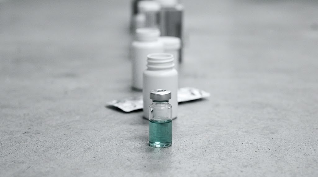 A glass vial of teal liquid in sharp focus at the front of a receding row of unlabeled white pill bottles under flat cold light