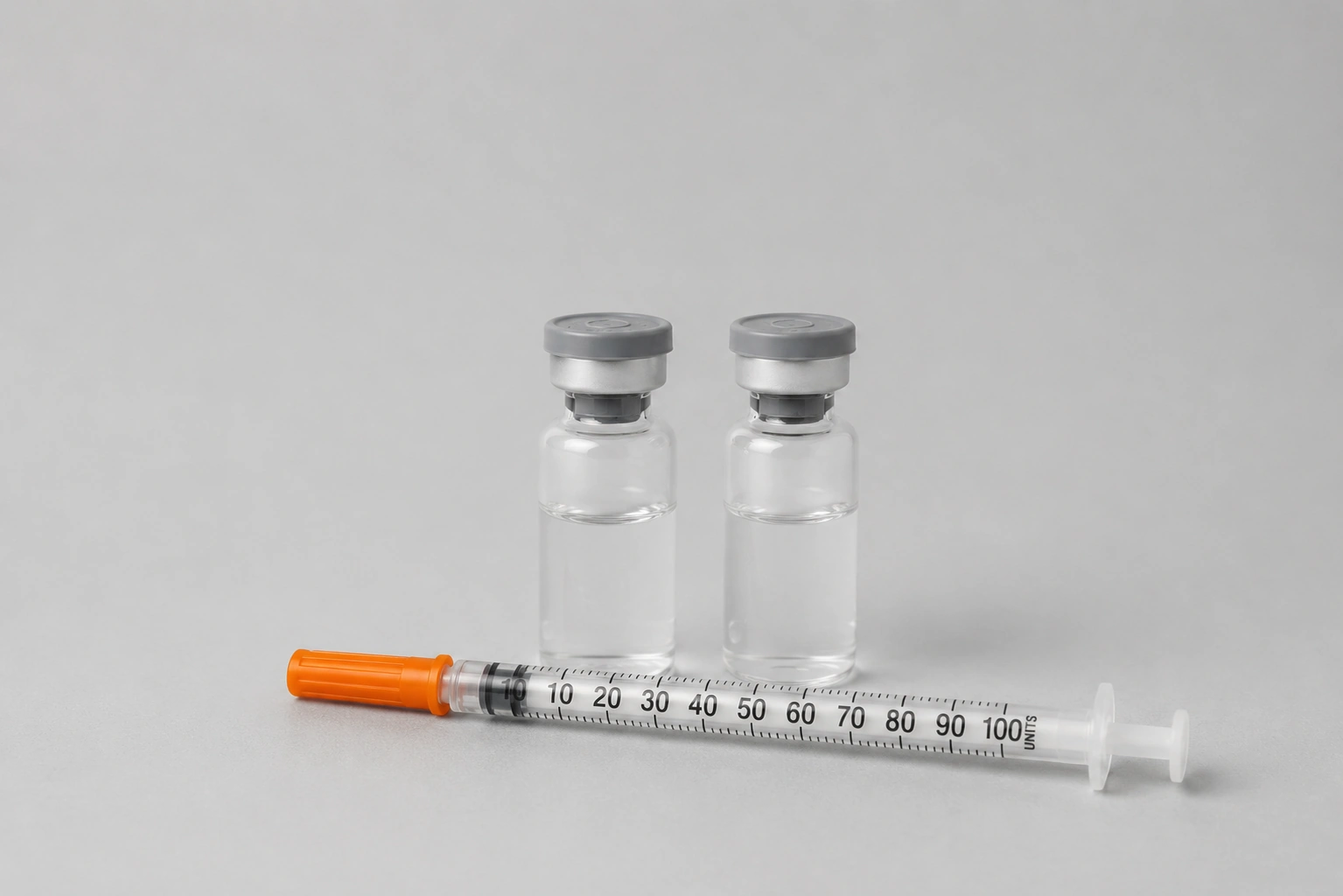 Two clear peptide vials with grey caps and a 1 mL U-100 insulin syringe with an orange plunger on a neutral background.