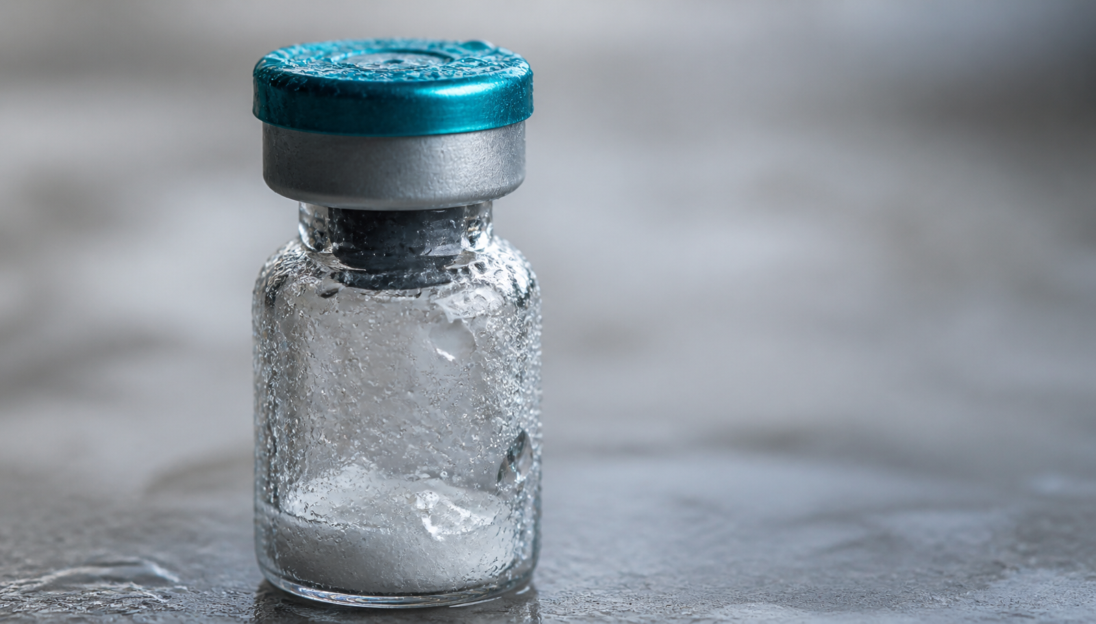 A sealed glass peptide vial with a teal cap on a gray surface, cold condensation beading across the glass.